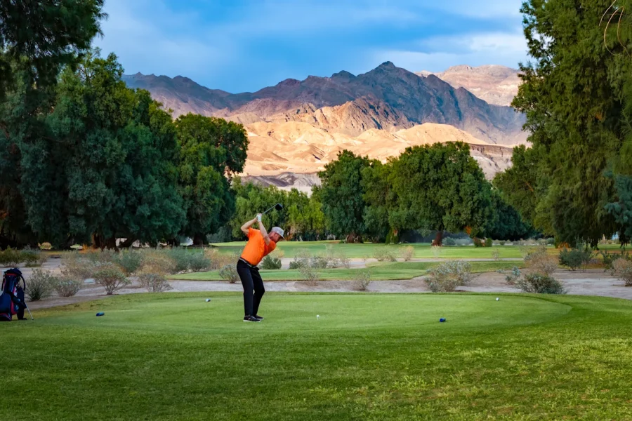 A golfer in an orange shirt is teeing off on a lush green course surrounded by trees, with mountains visible in the background under a clear blue sky. Golf clubs are in a bag on the ground nearby.