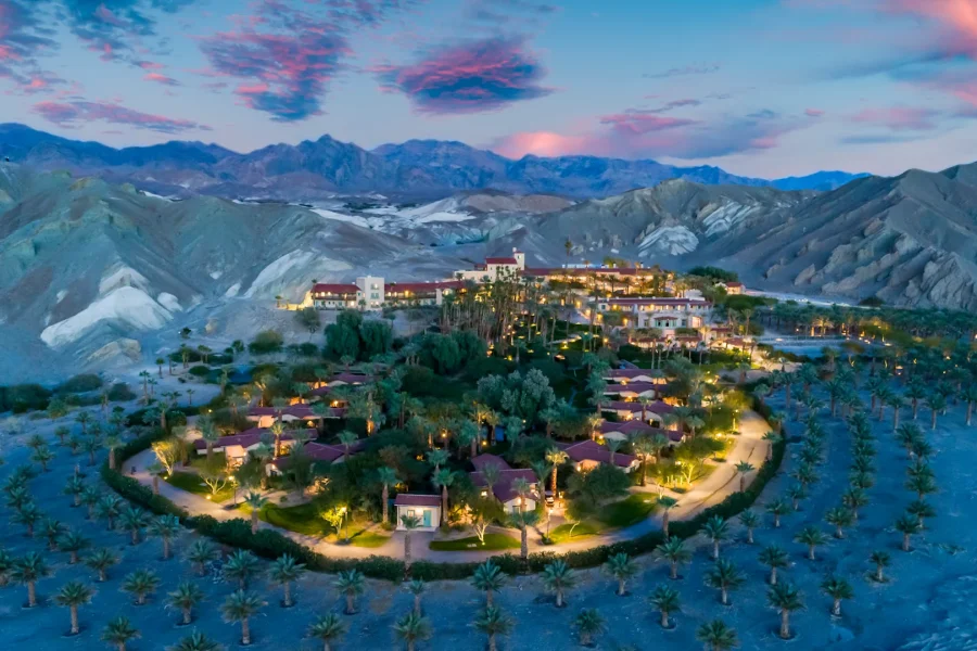 Aerial view of a circular resort with illuminated buildings and palm trees at dusk. Mountains and a vividly colored sunset sky form the backdrop, creating a serene desert oasis scene.
